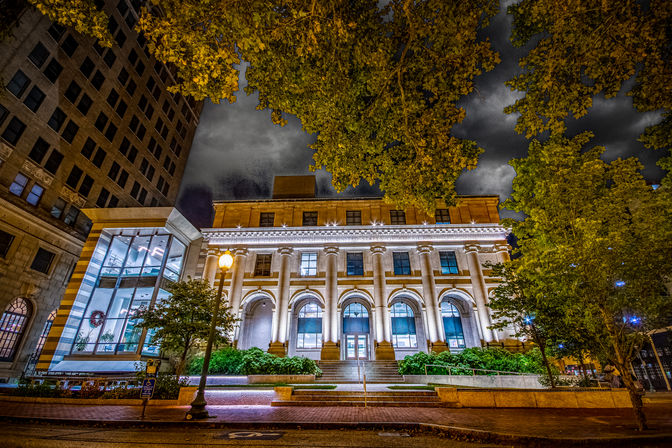 Night view of an illuminated neoclassical downtown building with grand columns and arched windows, a modern glass atrium to the left, leafy trees framing a dramatic cloudy sky.