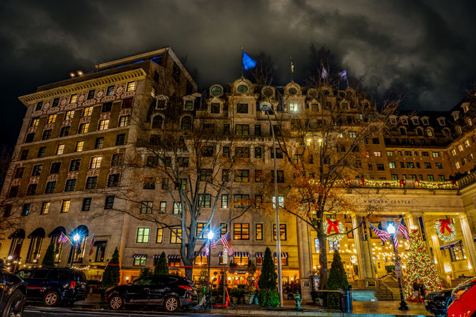 Historic downtown hotel facade at night with glowing holiday lights, wreaths and a decorated Christmas tree by the entrance, American flags, parked cars on a wet street and a dramatic cloudy sky.