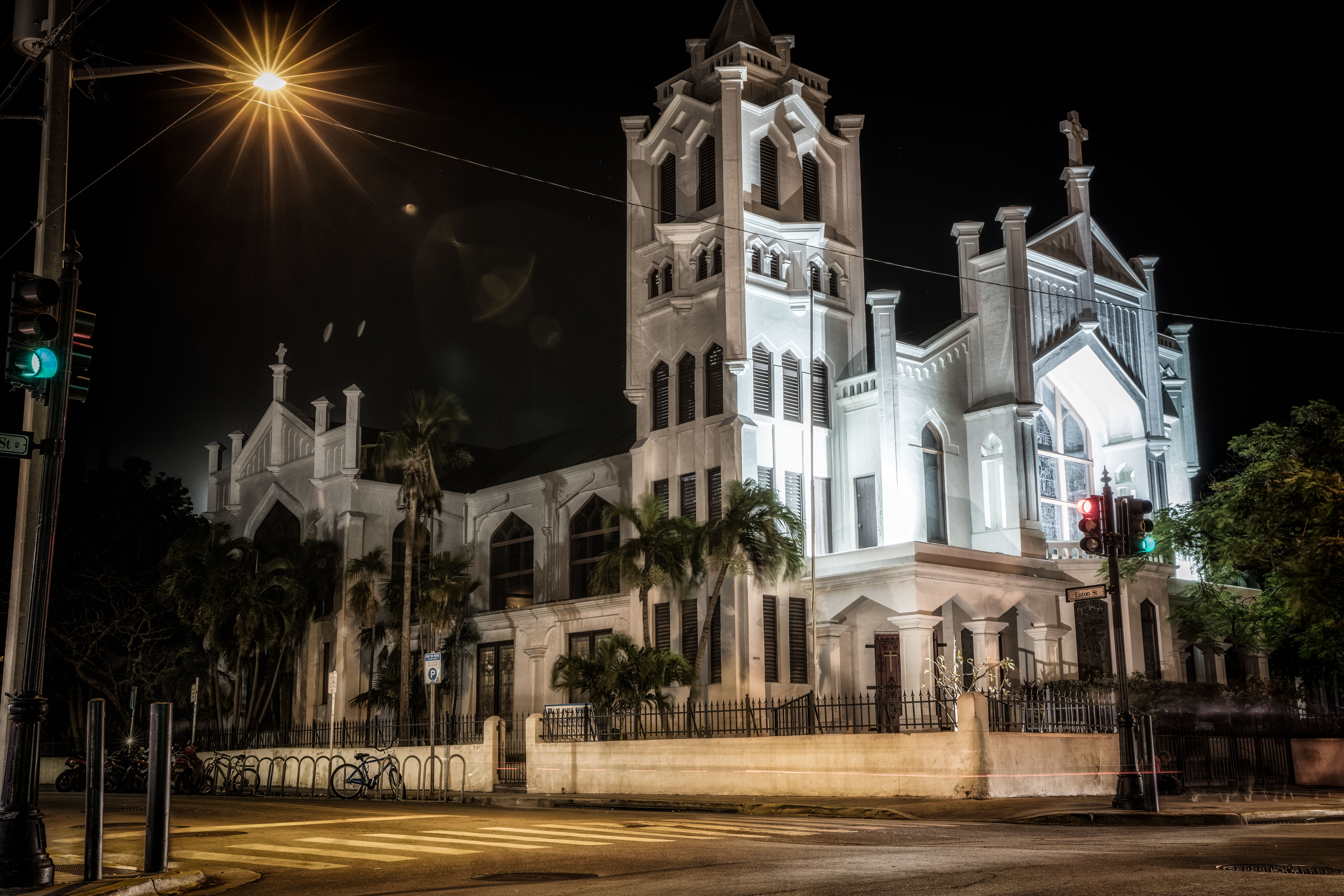 Illuminated historic Gothic church at night on an urban intersection, white stone facade and tower framed by palm trees, starburst streetlight and traffic signals