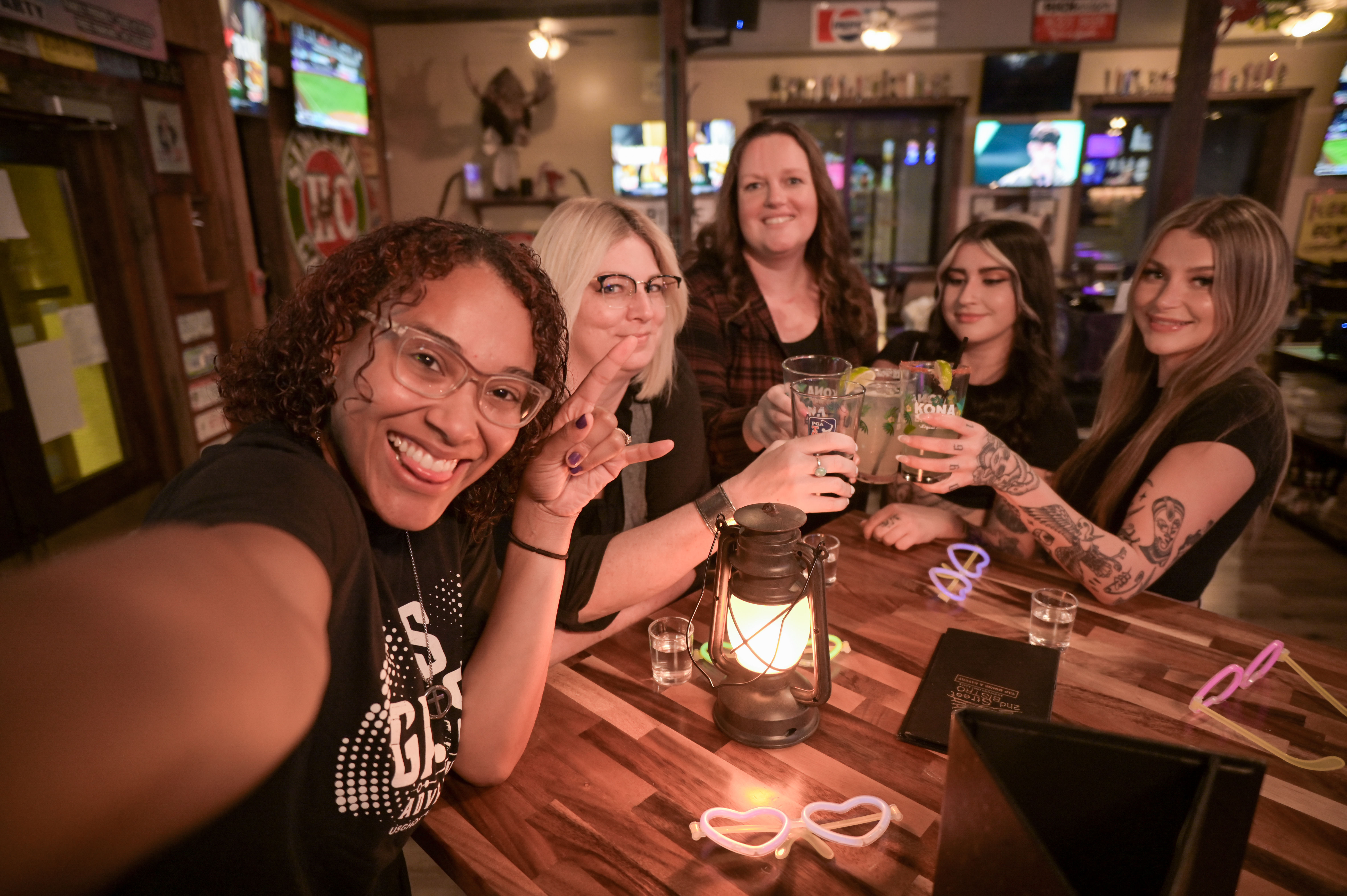 Selfie of five friends toasting with cocktails at a cozy neighborhood bar, wooden table with a glowing lantern, neon heart sunglasses on the table, visible tattoos and a lively evening vibe
