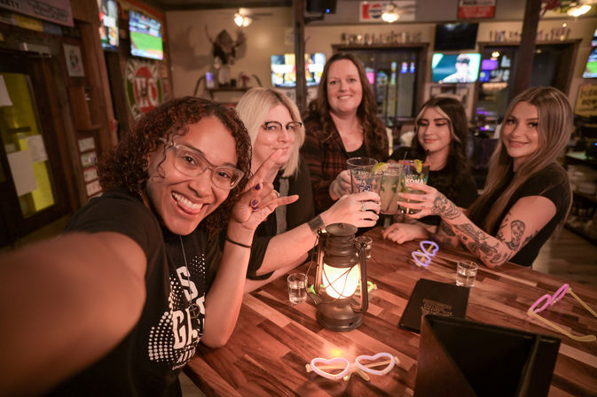 Selfie of five friends toasting with cocktails at a cozy neighborhood bar, wooden table with a glowing lantern, neon heart sunglasses on the table, visible tattoos and a lively evening vibe