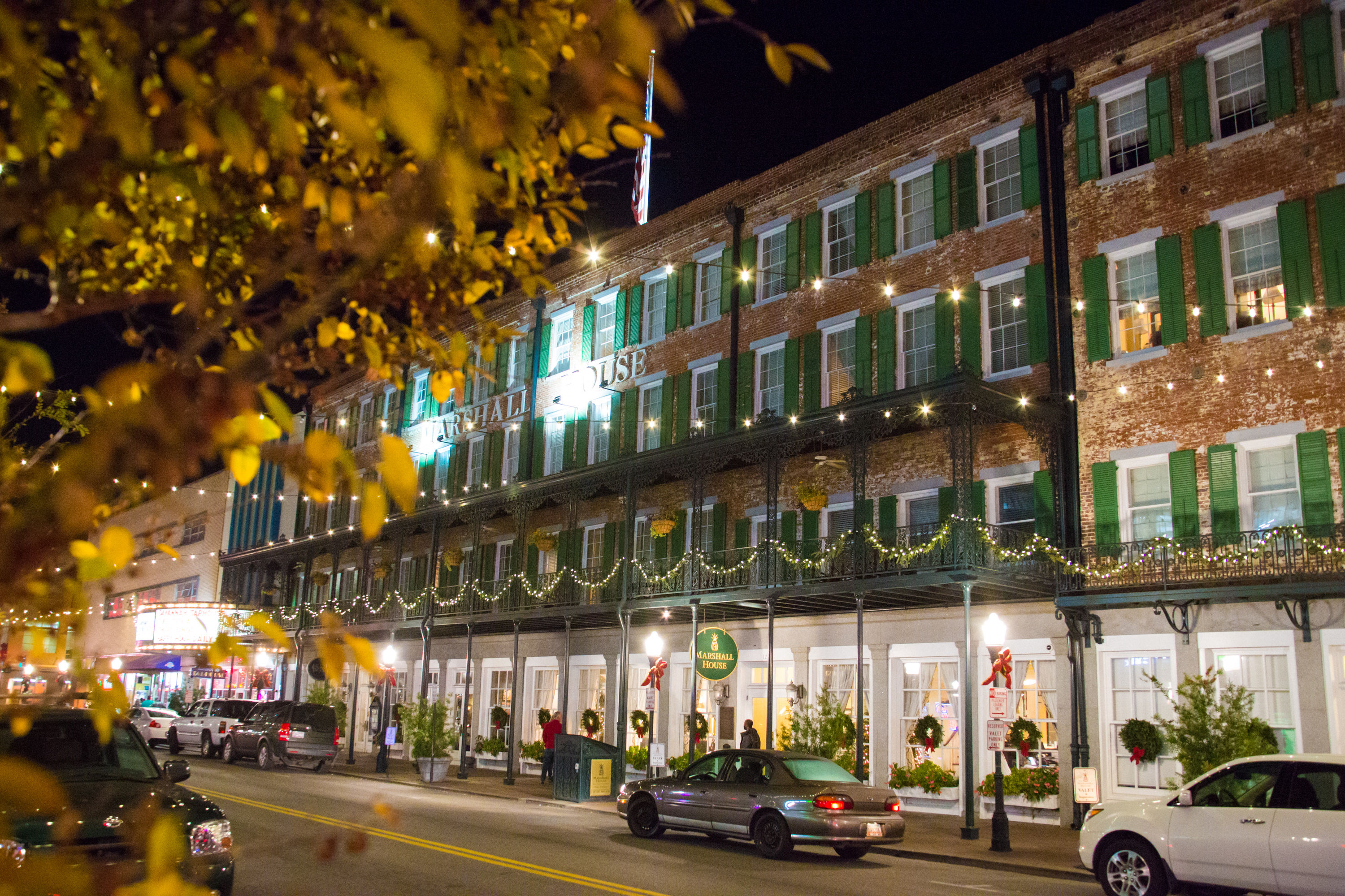 Nighttime downtown scene: historic three-story brick building with green shutters and iron balcony draped in festive string lights and wreaths, parked cars on the street and yellow-leaved tree in the foreground.