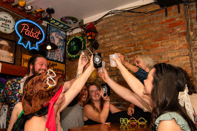 Smiling group of friends cheers with plastic cups and beer mugs in a cozy brick‑walled bar decorated with neon signage, vintage signs, novelty hats and glasses on the table.