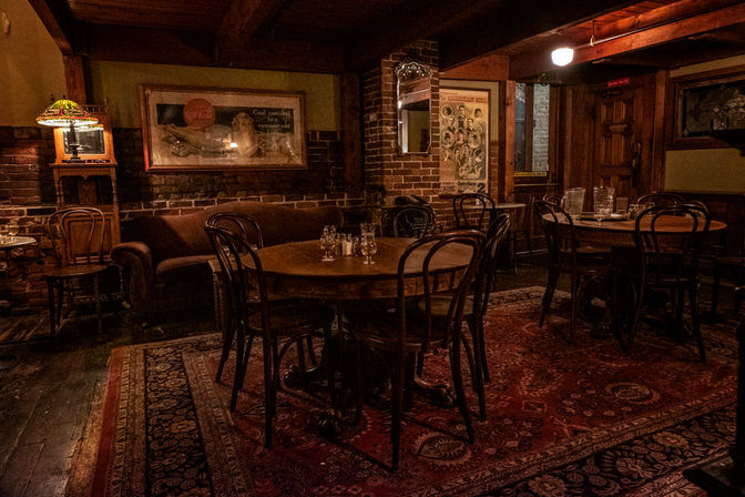 Cozy vintage pub interior with exposed brick walls, wooden round tables and bentwood chairs on a patterned rug, antique lamp and warm amber lighting.
