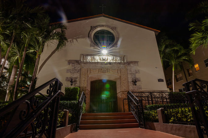 Night view of a Mediterranean-style chapel entrance with arched wooden doors, carved stone surround, wrought-iron railings and palm trees glowing under a bright exterior light.