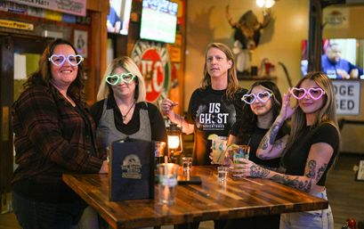 Five people at a wooden bar in a cozy pub wearing glowing heart-shaped glasses, holding drinks and a lantern — playful group night out.