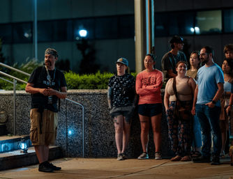 Nighttime downtown walking-tour scene: a bandana-wearing guide talks while a casual group of adults stands on a city sidewalk by steps, a railing and a lit building facade.