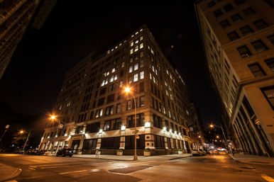 Downtown city street corner at night featuring a large historic multi-story brick building with glowing windows, amber streetlights, empty sidewalks and parked cars.