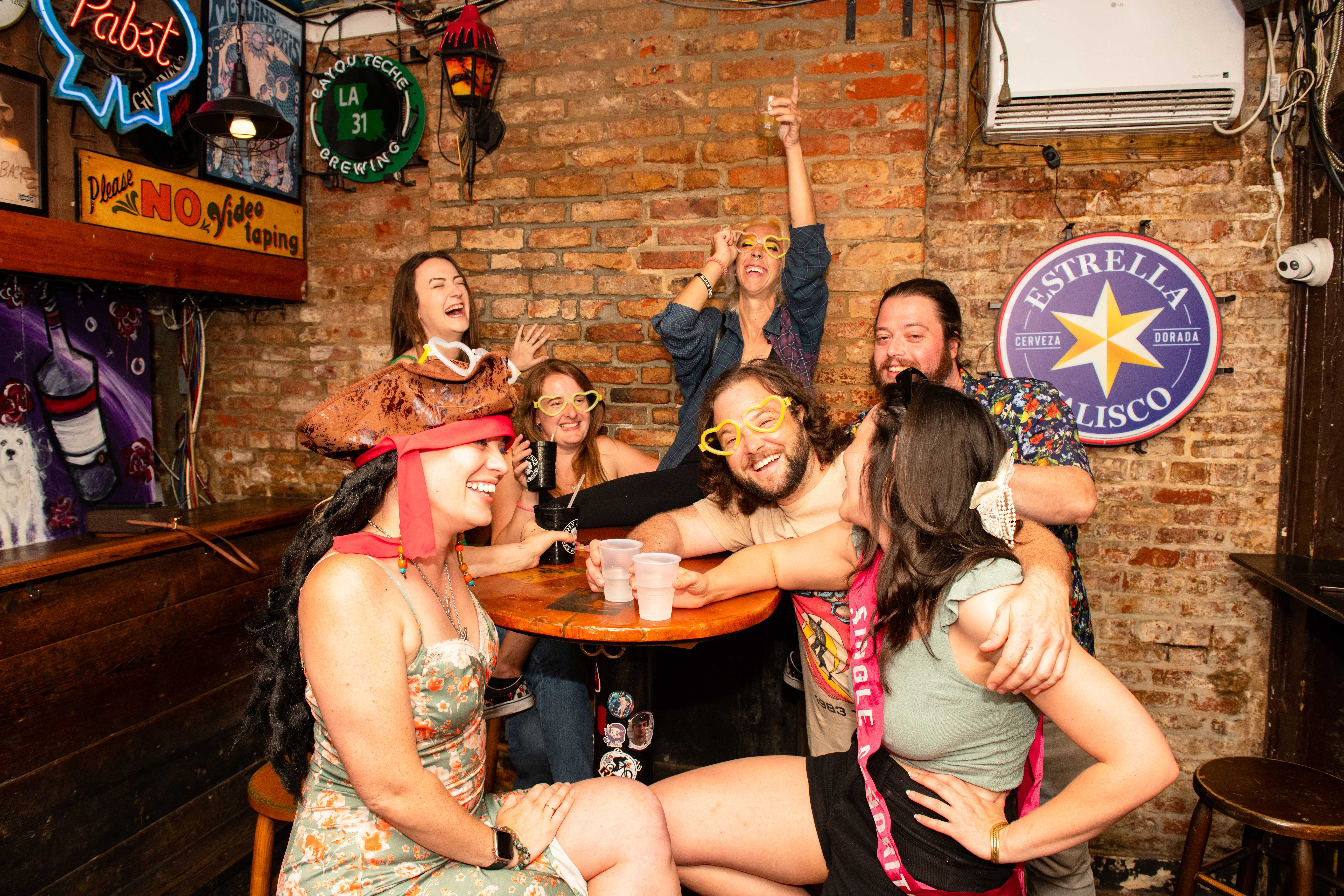 Seven friends in playful costumes and novelty glasses laughing and toasting around a wooden high-top in a cozy brick-walled neighborhood bar with neon signs and drinks.