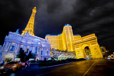 Nighttime Las Vegas Strip scene with glowing hotel tower, illuminated replica Eiffel Tower and triumphal arch, ornate casino façade and taxis under a dramatic cloudy sky