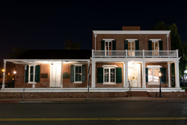 Historic two-story red-brick building with white columned porch, balcony and green shutters, warmly lit at night on a quiet main street with a vintage streetlamp.