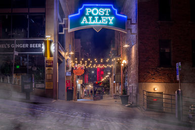 Neon-lit alleyway entrance at night with a glowing sign, overhead string lights, cobblestone street, brick buildings and soft fog creating a cozy urban nightlife scene.