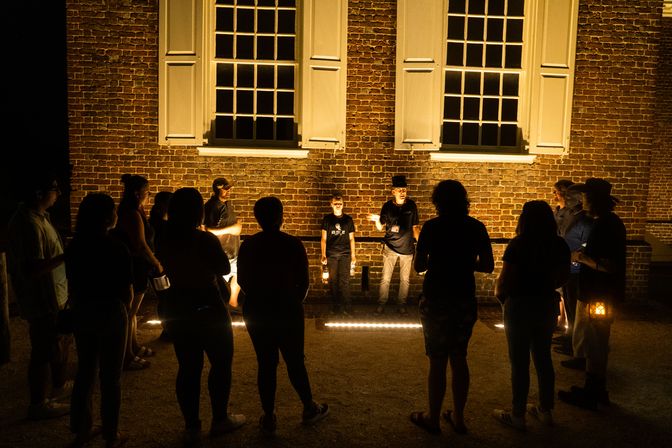 Nighttime lantern tour: visitors gathered in a semicircle outside a historic brick building with lit windows as a costumed guide gestures and tells stories.
