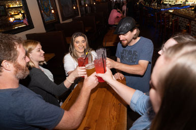 Six friends clinking colorful cocktails and a beer over a wooden table inside a dimly lit neighborhood bar, smiling and celebrating.