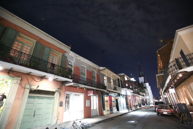 Nighttime scene in New Orleans French Quarter: narrow street lined with pastel two-story buildings and ornate wrought-iron balconies, lit storefronts, parked bicycles and cars, and a distant church steeple under a cloudy sky.