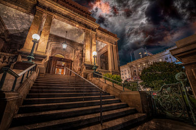 Grand stone steps leading to a historic neoclassical building entrance at night, glowing globe lampposts and ornate iron railings with downtown city buildings and dramatic storm clouds overhead.