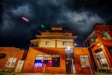 Nighttime downtown brick live-music bar with glowing neon OPEN sign, poster-covered windows, a parking meter out front, wooden upper floors and dramatic stormy clouds overhead.