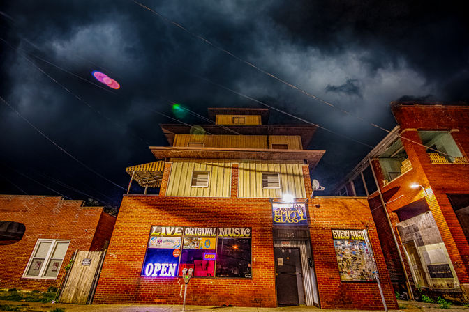 Nighttime downtown brick live-music bar with glowing neon OPEN sign, poster-covered windows, a parking meter out front, wooden upper floors and dramatic stormy clouds overhead.