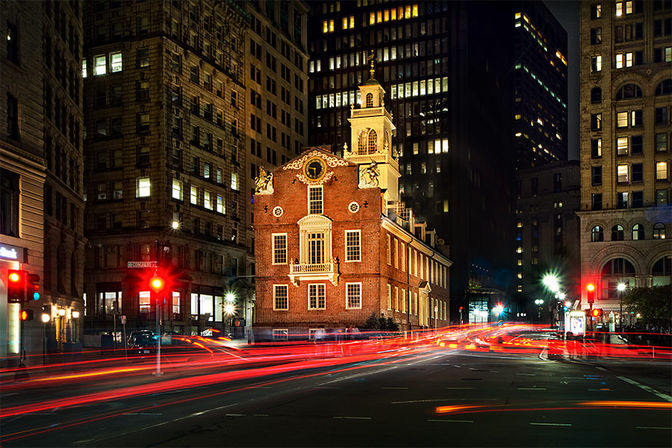 Historic red-brick colonial building glowing at night in a downtown intersection, framed by towering skyscrapers and streaking red and white car light trails.