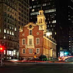 Nighttime view of a historic red-brick colonial building with a clock-topped cupola, brightly lit against modern skyscrapers, with streaking car light trails and traffic lights on a downtown city street.