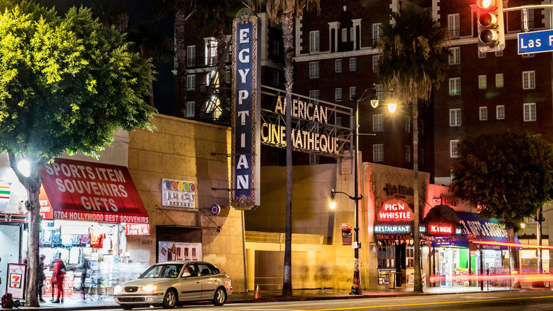Nighttime Hollywood, Los Angeles street with a lit historic movie-theater marquee, palm trees, souvenir shop awning, restaurants, a parked car and blurred pedestrians.
