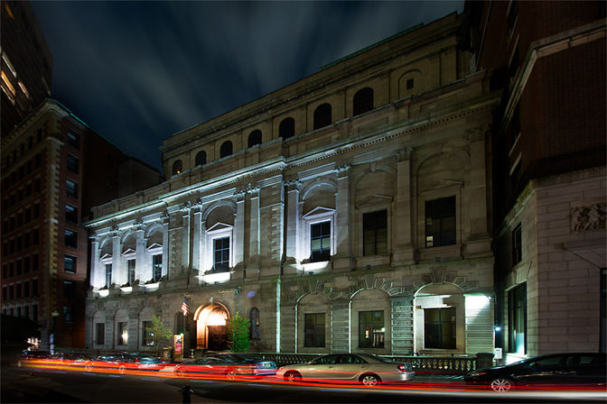 Night view of a historic neoclassical stone building on a downtown city street, floodlit arches and windows with parked cars and red light trails from passing traffic.
