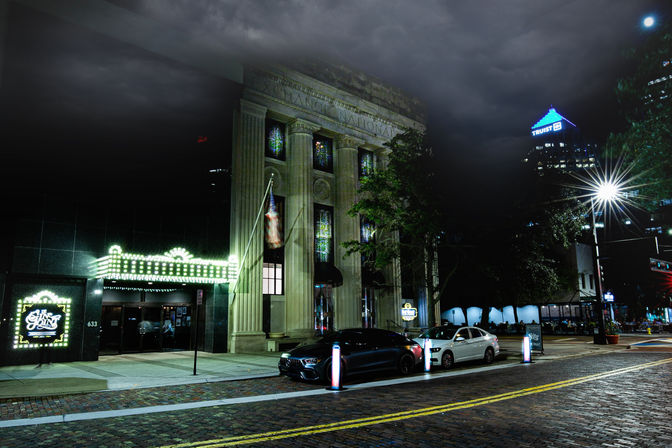 Downtown night scene with a neoclassical columned building and glowing marquee, brick-paved street with parked cars, bright streetlight and a lit city tower under moody clouds.