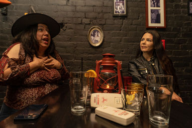 Two women chatting at a dim brick-walled bar table with a red lantern centerpiece, empty pint and cocktail glasses with lemon slices, a smartphone on the table and framed photos on the wall — lively night out scene.