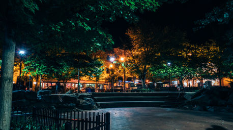 Nighttime urban park plaza with glowing streetlights, leafy trees, stone steps and benches in front of a row of warmly lit storefronts.