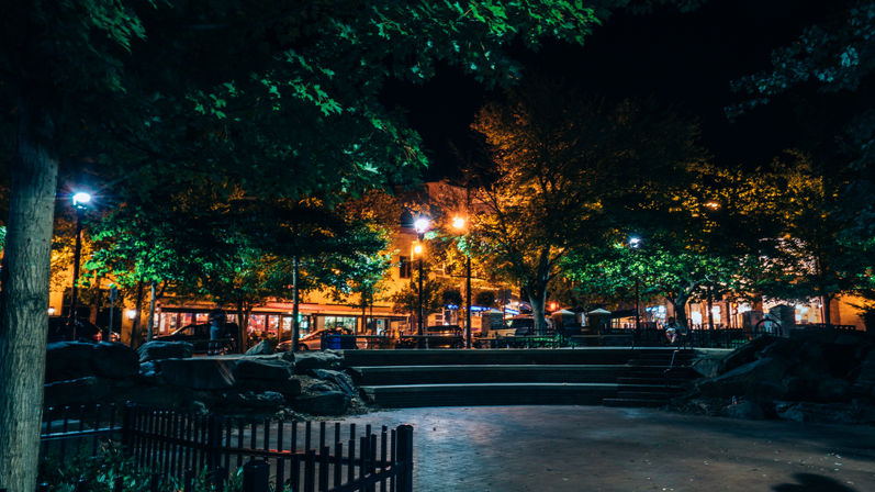 Nighttime urban park plaza with glowing streetlights, leafy trees, stone steps and benches in front of a row of warmly lit storefronts.