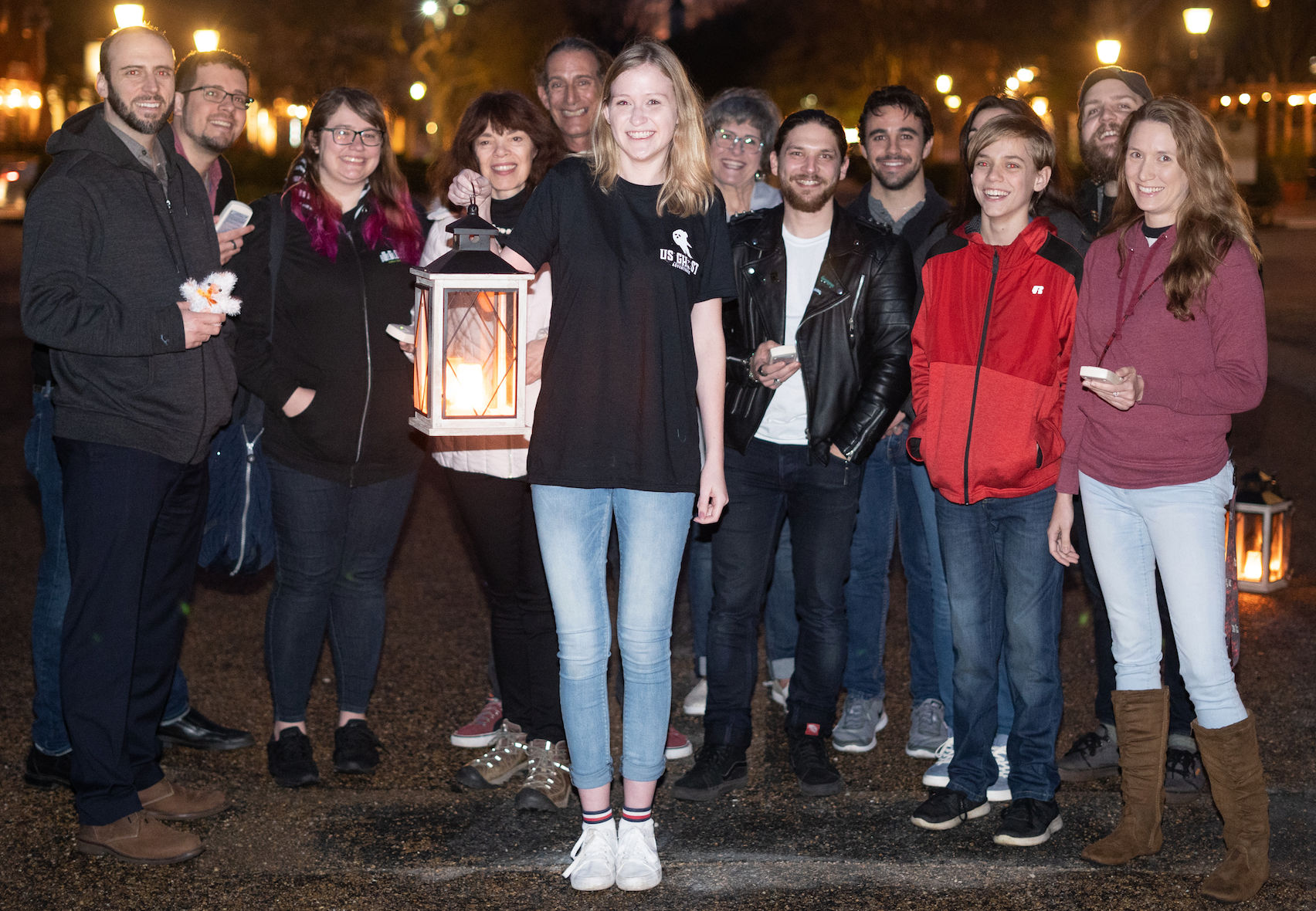 Smiling community group on a city street at night, a young woman at center holding a glowing lantern during a casual evening lantern walk.