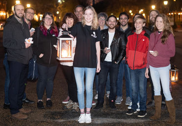 Smiling community group on a city street at night, a young woman at center holding a glowing lantern during a casual evening lantern walk.