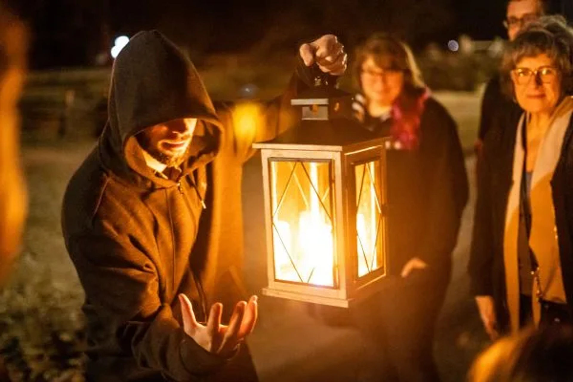 Hooded person theatrically holding a glowing lantern at an outdoor nighttime gathering, warm light illuminating smiling onlookers