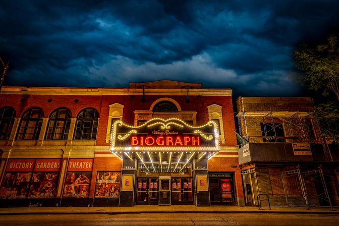Glowing vintage theater marquee on a red-brick downtown theater at night, dramatic storm clouds overhead and an empty city street below.