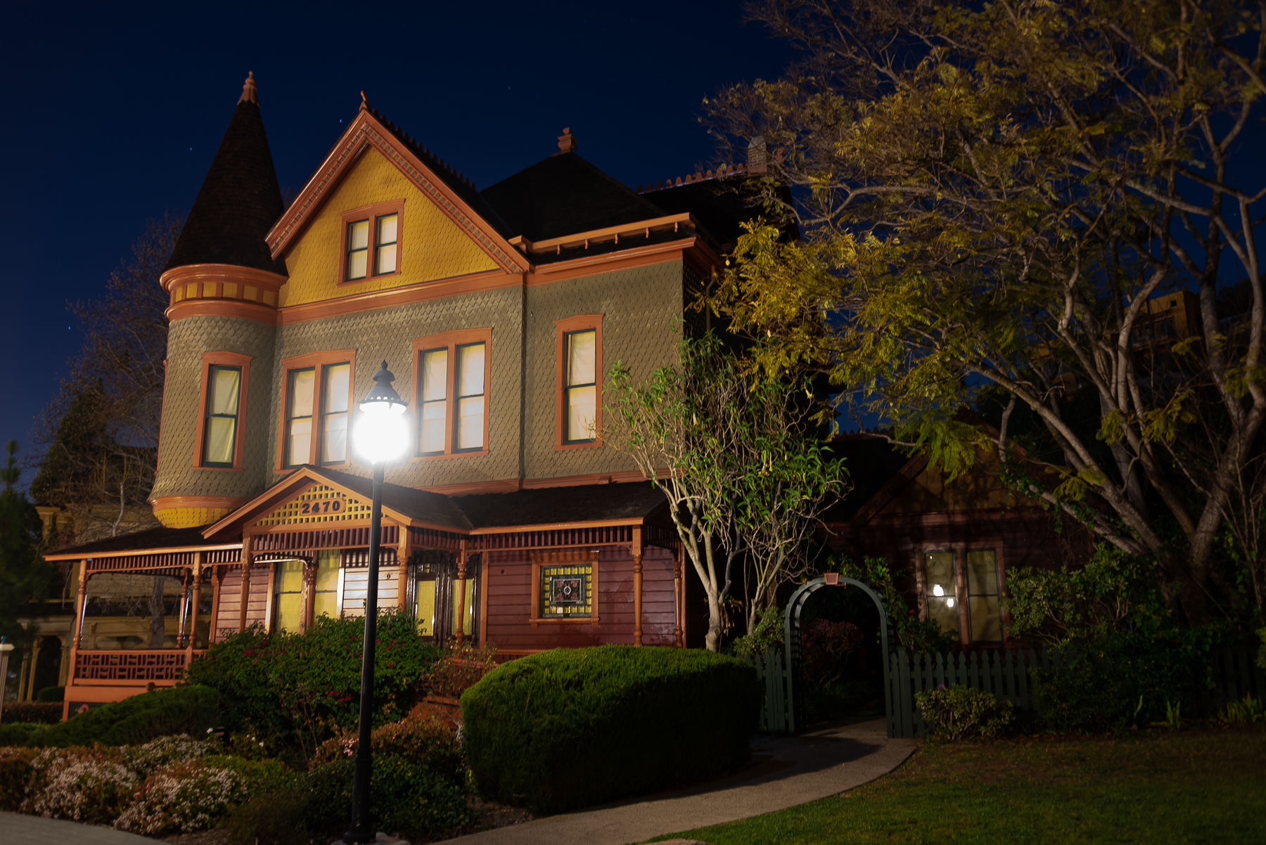 Victorian house at night with a turret and ornate porch, glowing streetlamp and garden arch in a leafy residential neighborhood