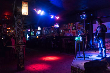 Moody live-music bar interior at night with a solo guitarist on a small stage under blue and pink spotlights, a red-lit dance floor, patrons at the dim bar, and sticker-covered walls and pillars.