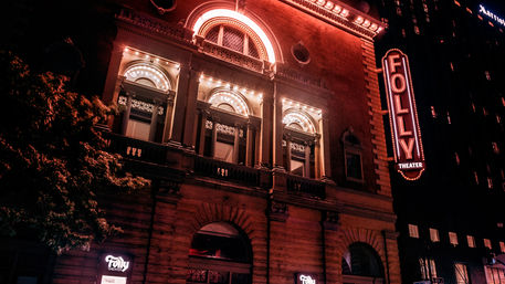 Nighttime urban scene of a historic brick theater facade with ornate arched windows, glowing marquee and vertical neon sign casting warm red light over the downtown street.