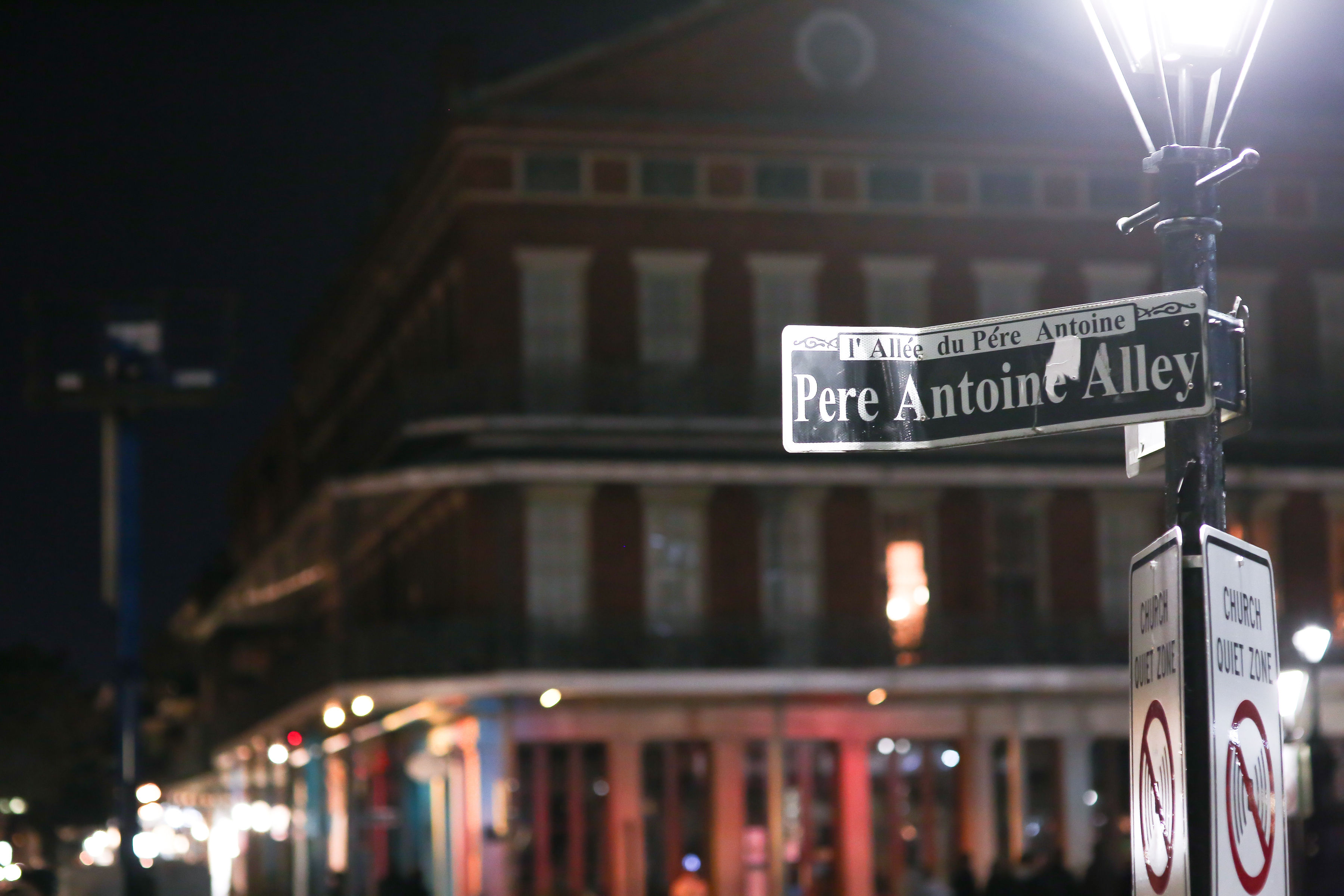 Illuminated "Pere Antoine Alley" street sign on a lamppost at night, with a blurred historic brick building and colorful storefronts in the background.