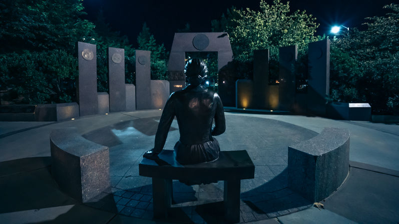 Bronze statue seated on a bench in a circular stone memorial plaza at night, moody blue lighting highlighting vertical stone pillars and surrounding trees