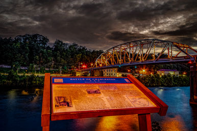 Illuminated historical marker titled "Battle of Charleston" overlooking a river at dusk with a lit steel-truss bridge, glowing city lights, and a dramatic cloudy sky.