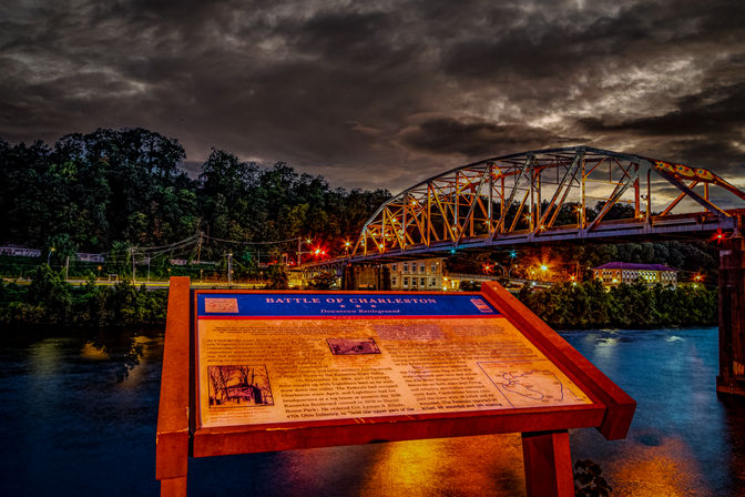 Illuminated historical marker titled "Battle of Charleston" overlooking a river at dusk with a lit steel-truss bridge, glowing city lights, and a dramatic cloudy sky.