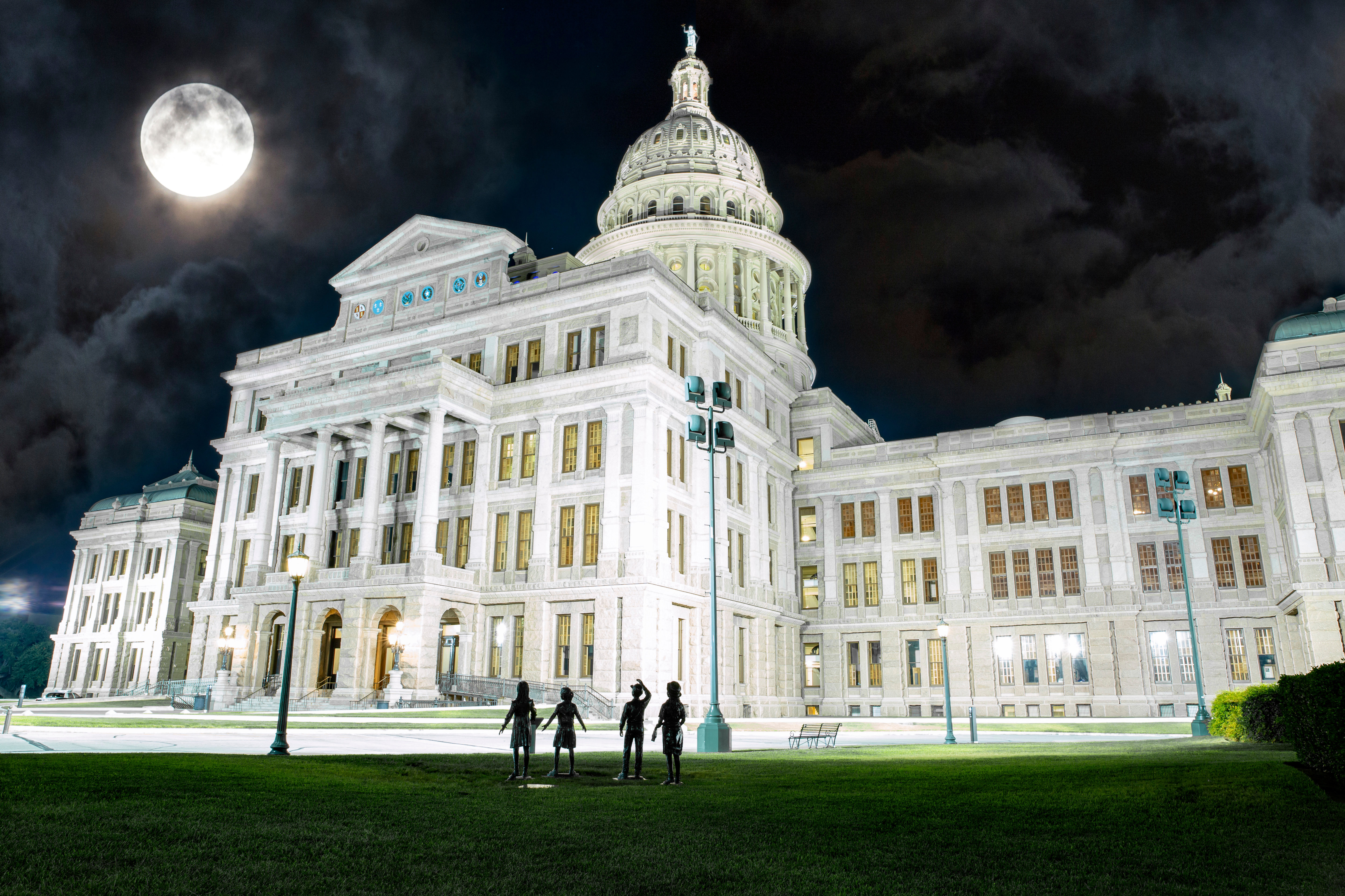 Illuminated neoclassical state capitol building at night with a glowing full moon, dramatic clouds, and four silhouetted figures on the front lawn.