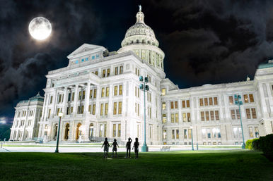 Illuminated neoclassical state capitol building at night with a glowing full moon, dramatic clouds, and four silhouetted figures on the front lawn.