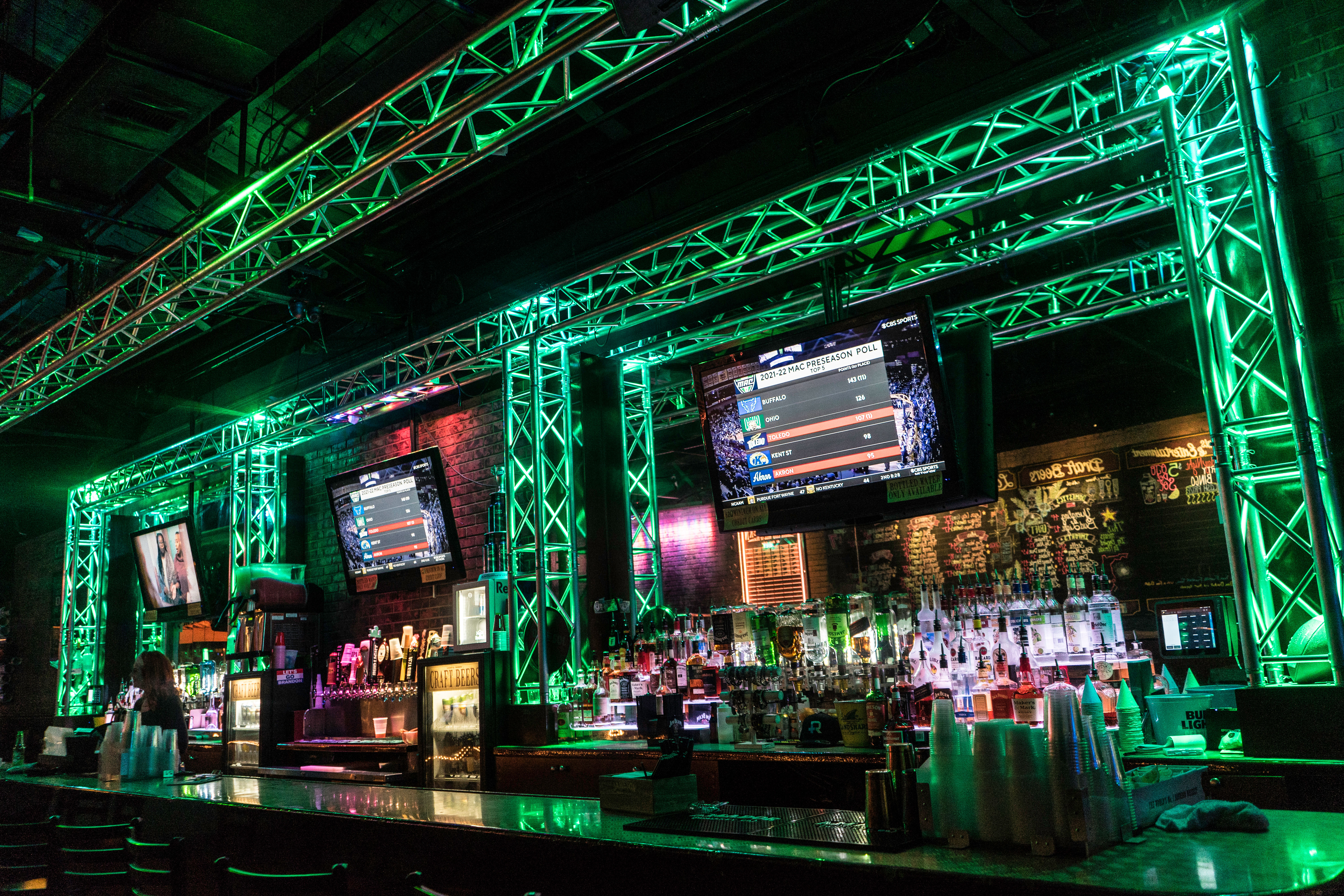 Neon-green lit sports bar interior with metal truss framing, rows of liquor bottles and taps behind a glossy bar counter, multiple TV screens showing scores, stacked plastic cups and bar stools — vibrant urban nightlife vibe.