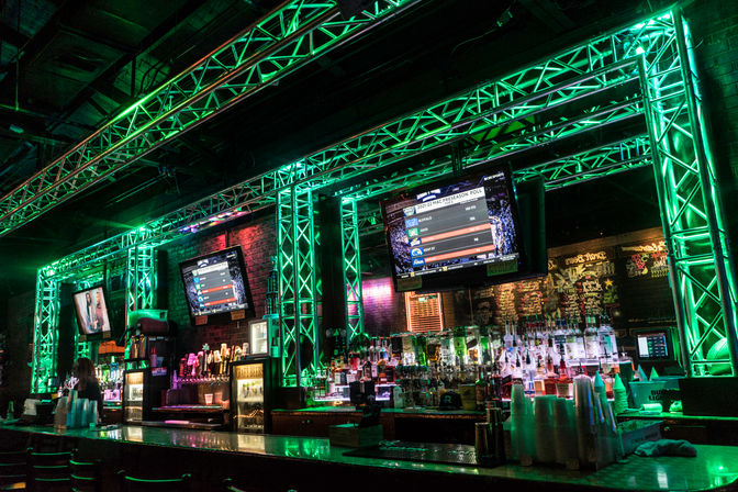 Neon-green lit sports bar interior with metal truss framing, rows of liquor bottles and taps behind a glossy bar counter, multiple TV screens showing scores, stacked plastic cups and bar stools — vibrant urban nightlife vibe.