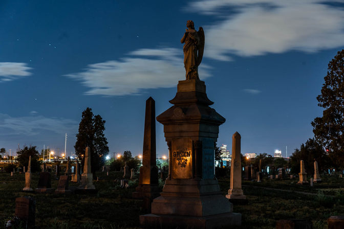 Night cemetery with an angel-topped stone monument and obelisks in the foreground, scattered gravestones, distant city lights and clouds under a starry sky.