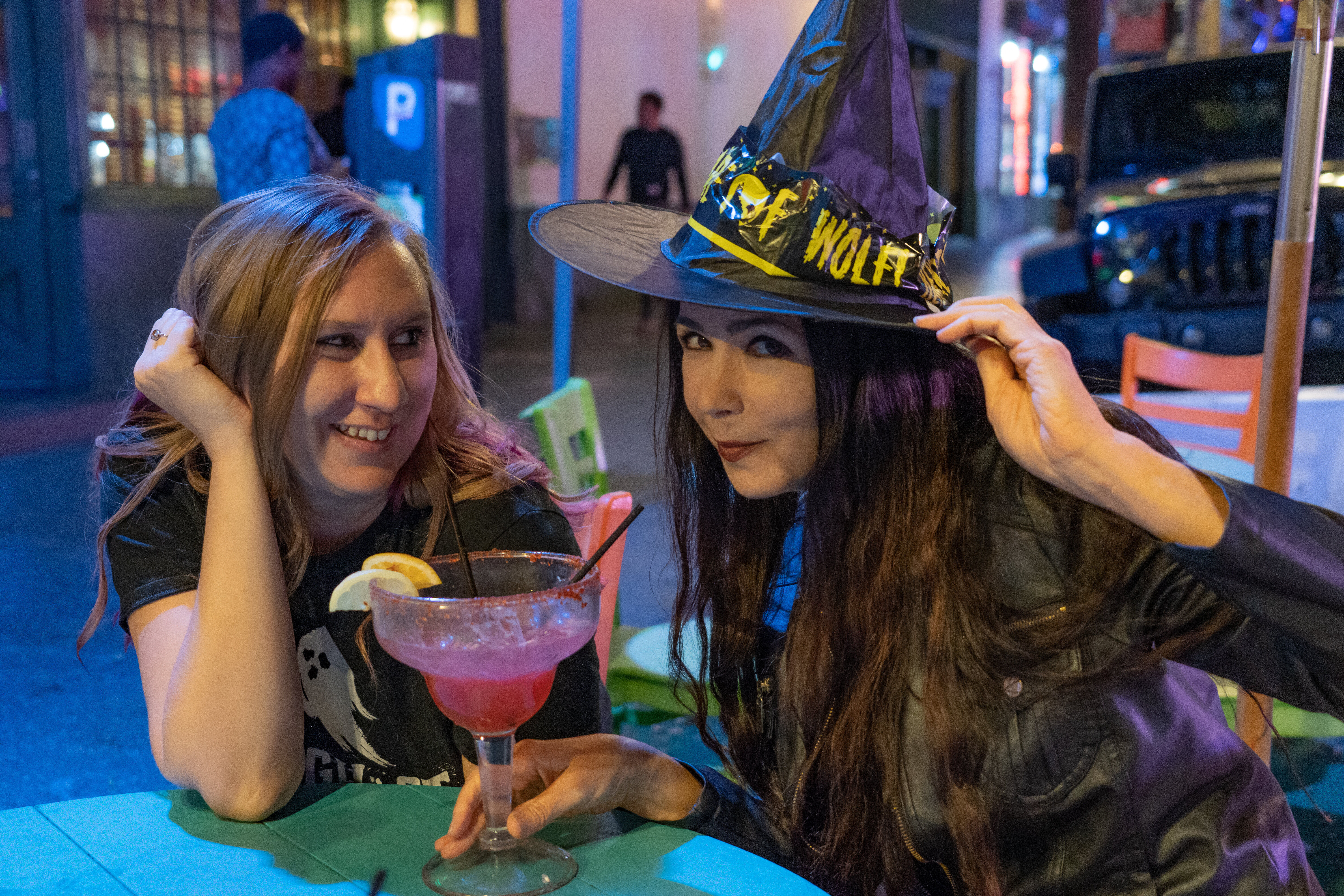 Two friends enjoying downtown nightlife on an outdoor patio at night — one playfully tipping a Halloween witch hat, the other smiling as they share a large pink cocktail with lemon garnish under neon lights.