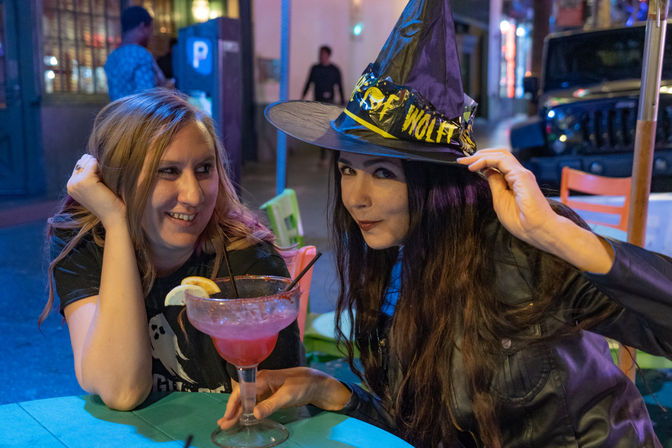 Two friends enjoying downtown nightlife on an outdoor patio at night — one playfully tipping a Halloween witch hat, the other smiling as they share a large pink cocktail with lemon garnish under neon lights.