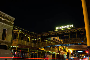 Historic downtown at night: illuminated covered pedestrian bridge above ornate two-story brick buildings with warm string lights and streaking car lights.