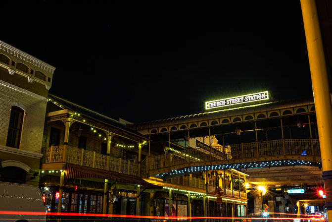 Historic downtown at night: illuminated covered pedestrian bridge above ornate two-story brick buildings with warm string lights and streaking car lights.